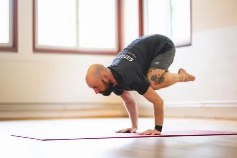 Man practicing crow pose yoga on a red mat in a studio.