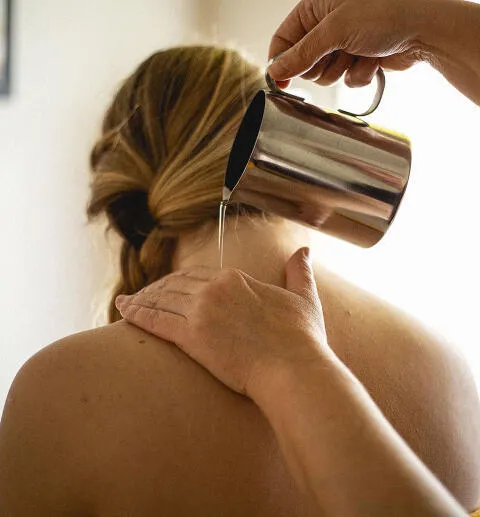 A person pouring oil or water from a metal pitcher onto a woman's shoulder during a spa treatment.