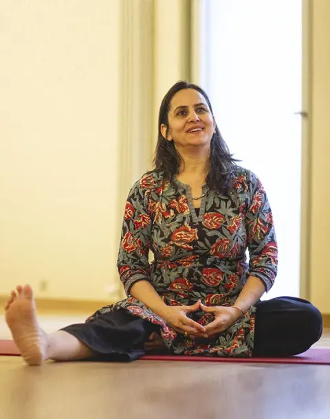 A woman in a floral tunic sitting on a red yoga mat with one leg extended, smiling and looking upward.