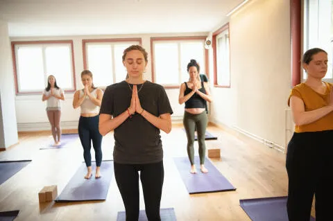 A group of women in a bright studio standing on purple mats with eyes closed and hands in prayer position (Anjali Mudra) during a yoga class.