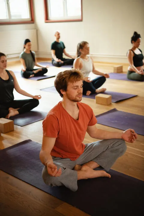 A group of people sitting in cross-legged meditation pose (Sukhasana) with eyes closed and hands resting on their knees during a yoga class.