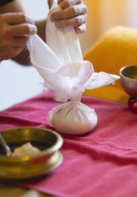 Hands tying a white cloth bundle over a red fabric surface with brass bowls nearby.
