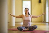 A smiling woman in a pink tank top and patterned leggings sitting cross-legged on a red yoga mat with her arms outstretched in a bright, open studio.