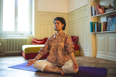A woman in a floral tunic sitting cross-legged in a meditative pose on a purple yoga mat in a cozy, sunlit room with bookshelves.
