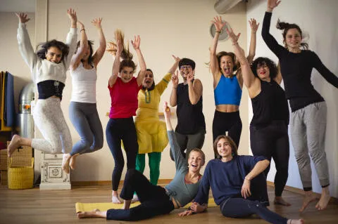 A diverse and joyful group of women celebrating in a yoga studio, with several participants jumping in the air and others smiling on the floor.