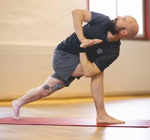 A man practices yoga in a studio, performing a twisted prayer lunge on a red mat.