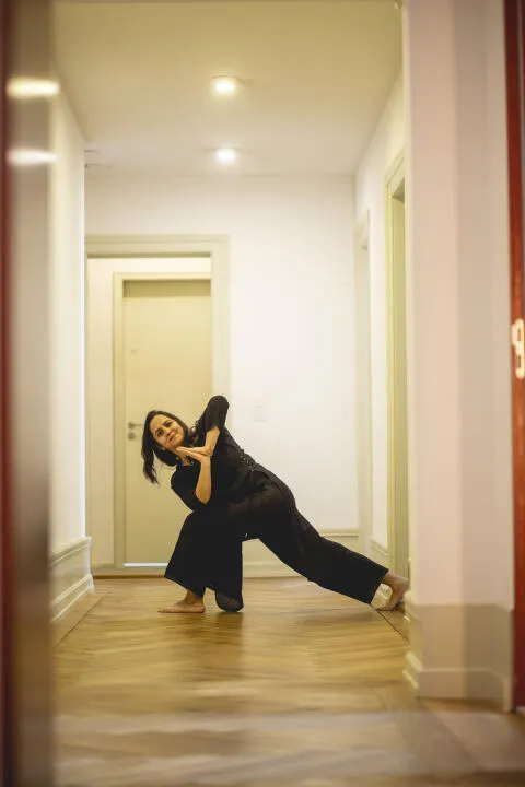 A woman in black clothing performing a Revolved Side Angle yoga pose (Parivrtta Parsvakonasana) on a wooden floor in a brightly lit white hallway.