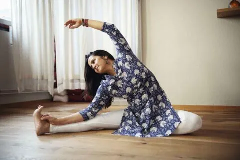 A woman in a blue floral tunic and white leggings performing a seated side stretch yoga pose on a wooden floor.