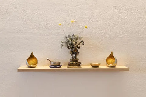 A wooden floating shelf featuring a bronze Ganesha statue, a Tibetan singing bowl, and lit tea light candles against a textured wall.