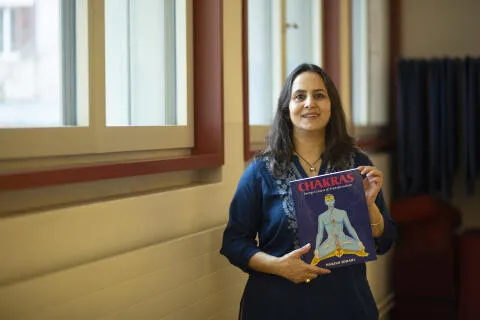 A smiling woman in a blue tunic holding the book 'Chakras: Energy Centers of Transformation' by Harish Johari in a bright studio.