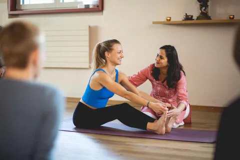 A smiling yoga teacher in a pink tunic guiding a student in a blue top through a seated forward fold stretch in a bright studio.