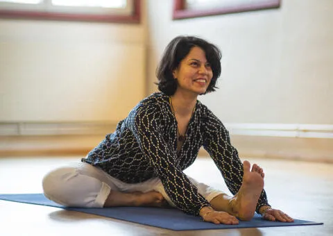 Smiling woman in a patterned tunic performing a head-to-knee forward bend (Janu Sirsasana) on a blue yoga mat.