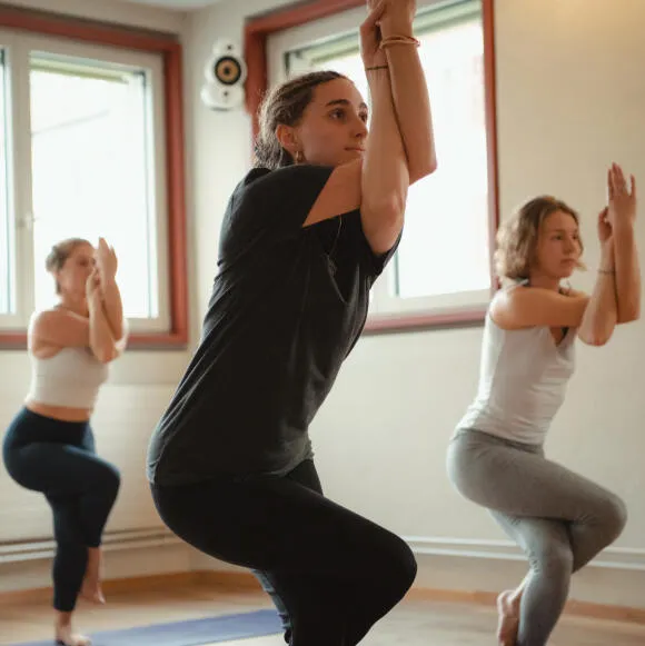 Three women in a yoga studio practicing Eagle Pose (Garudasana), focusing on their balance with arms and legs entwined.
