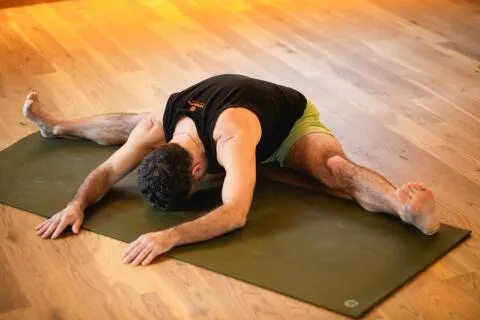 A man performing a seated wide-legged forward fold yoga pose with arms extended on a green mat.