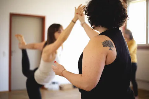 A yoga instructor with a tree tattoo on her shoulder guiding a student in a standing dancer's pose (Natarajasana).