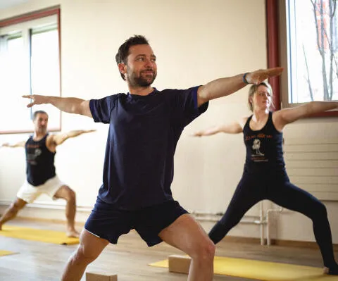 Three yoga practitioners performing Warrior II (Virabhadrasana II) on yellow mats in a bright studio.
