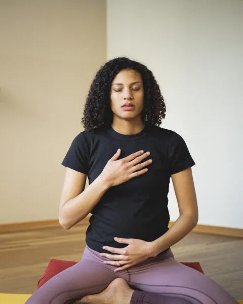 Woman with curly hair meditating in a seated yoga pose, with one hand on her chest and the other on her abdomen.