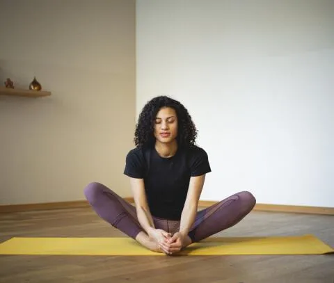 Woman with curly hair sitting in a butterfly yoga pose with eyes closed on a yellow mat.