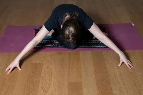 Woman performing a seated forward fold yoga pose with arms extended on a yoga mat.