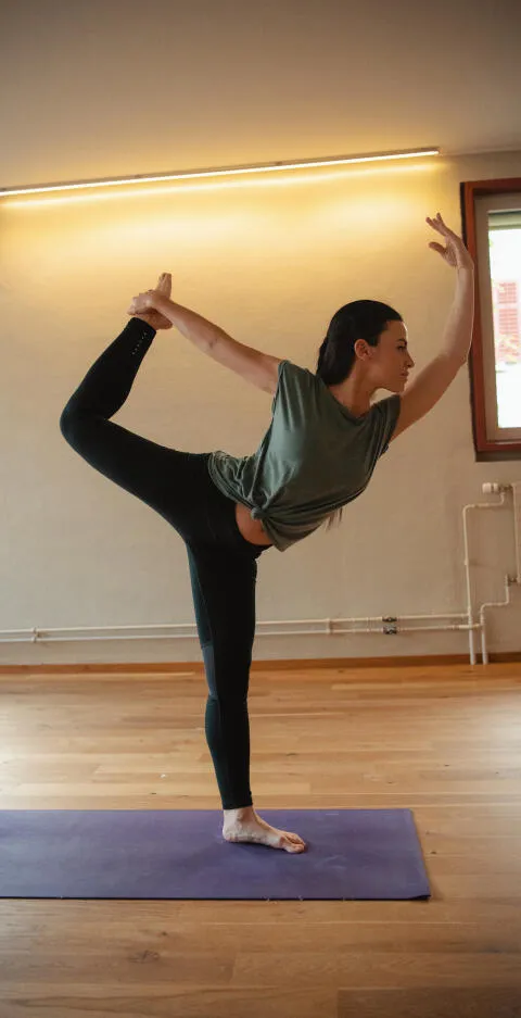 Woman in a green t-shirt and black leggings performing a standing dancer's pose (Natarajasana) on a purple mat in a studio.