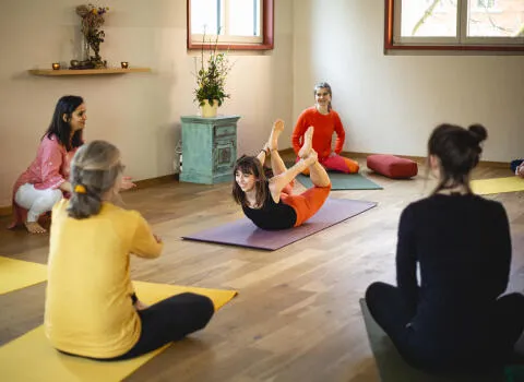 A woman in a black tank top and orange leggings performing bow pose (Dhanurasana) in a bright yoga studio surrounded by students.