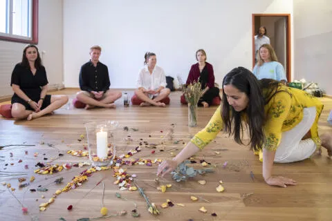 A woman in a yellow tunic arranging a flower mandala on a wooden floor during a meditation circle with participants sitting in the background.