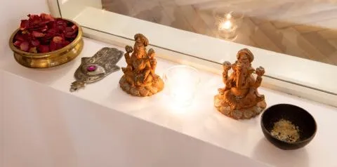 A peaceful spiritual display with small wooden statues, rose petals, and a glowing candle set against a window.