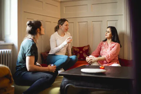 Three women sitting comfortably on cushions in a cozy corner, drinking tea and having a relaxed conversation in a yoga studio setting.