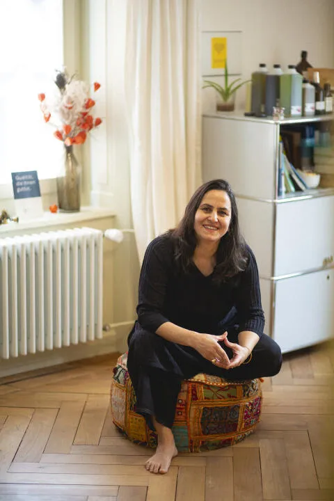 Smiling woman in black attire sitting cross-legged on a colorful embroidered floor pouf in a bright, modern wellness studio.