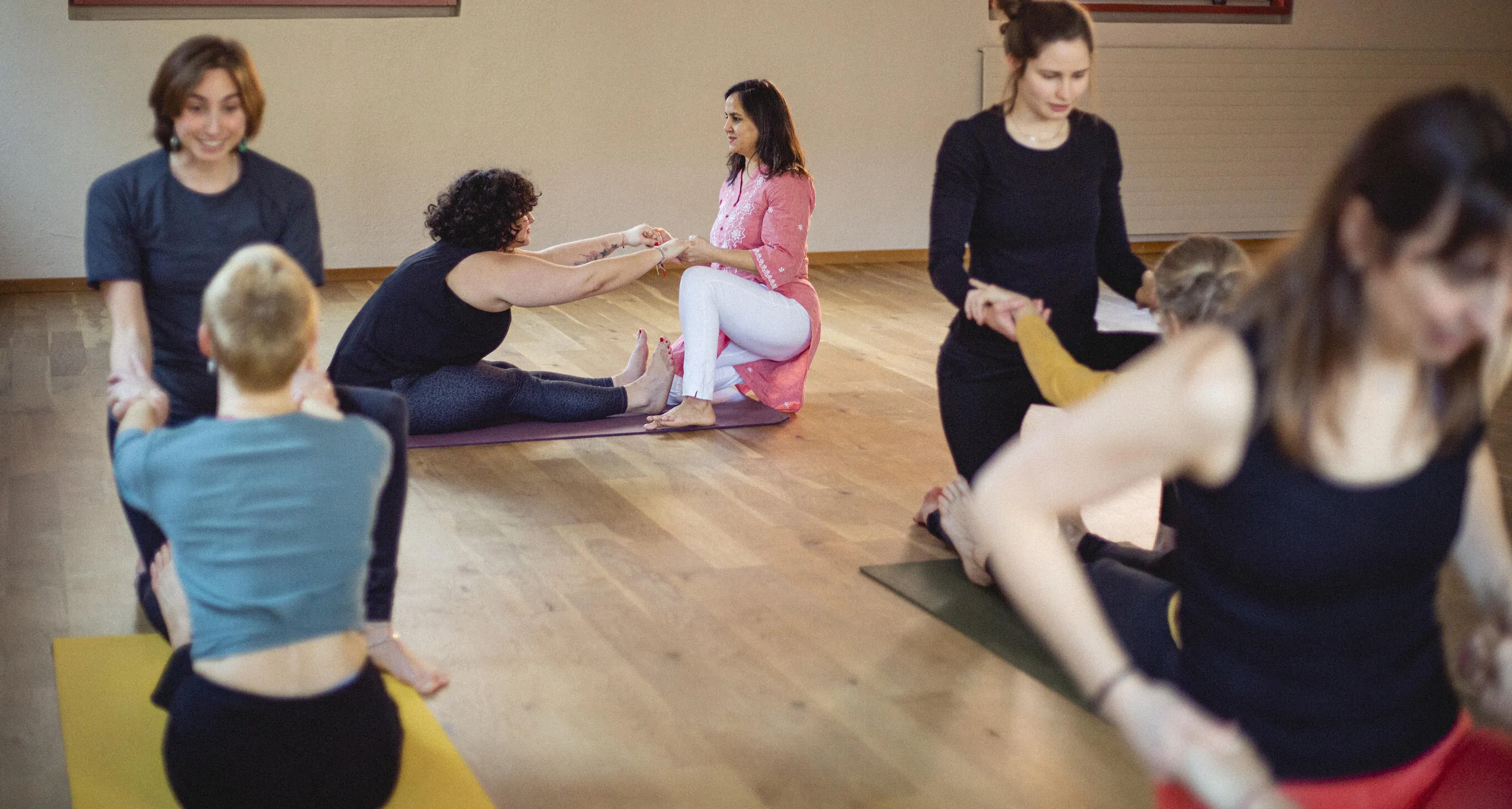 People practicing partner yoga poses in a spacious wooden floor studio.